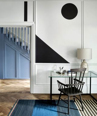 White painted office with black geometric shapes painted on wall, glass desk, wooden chair, table lamp, wooden floor, blue rug, blue painted stairs in background in hallway