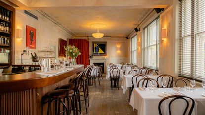 A modern bistro with wooden floors, chairs, and bar counter, colorful figurative artworks on distressed, rose-tinted cement walls, red curtains, white table cloths, and golden-lit sconces.