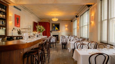 A modern bistro with wooden floors, chairs, and bar counter, colorful figurative artworks on distressed, rose-tinted cement walls, red curtains, white table cloths, and golden-lit sconces.