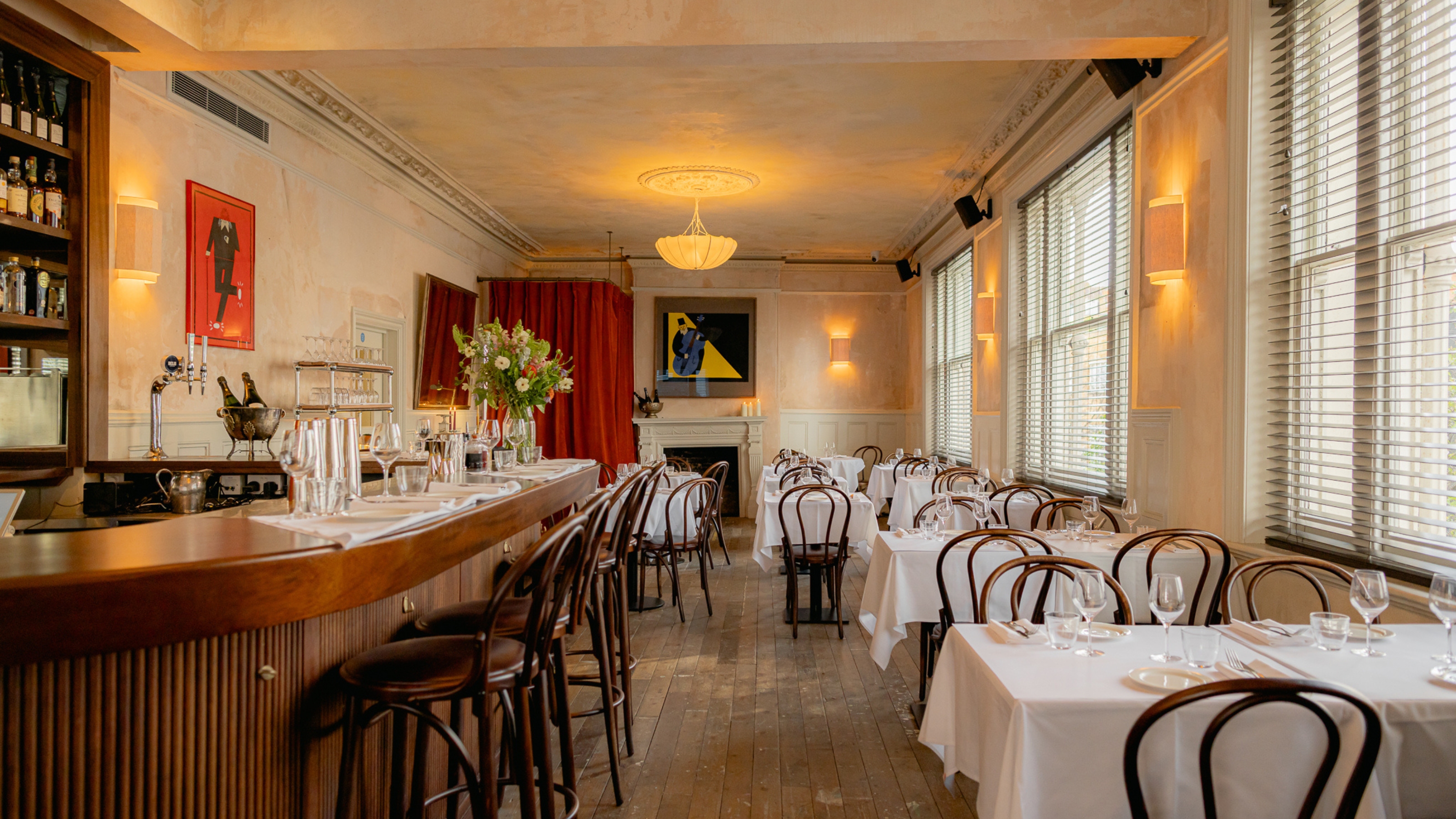 A modern bistro with wooden floors, chairs, and bar counter, colorful figurative artworks on distressed, rose-tinted cement walls, red curtains, white table cloths, and golden-lit sconces.