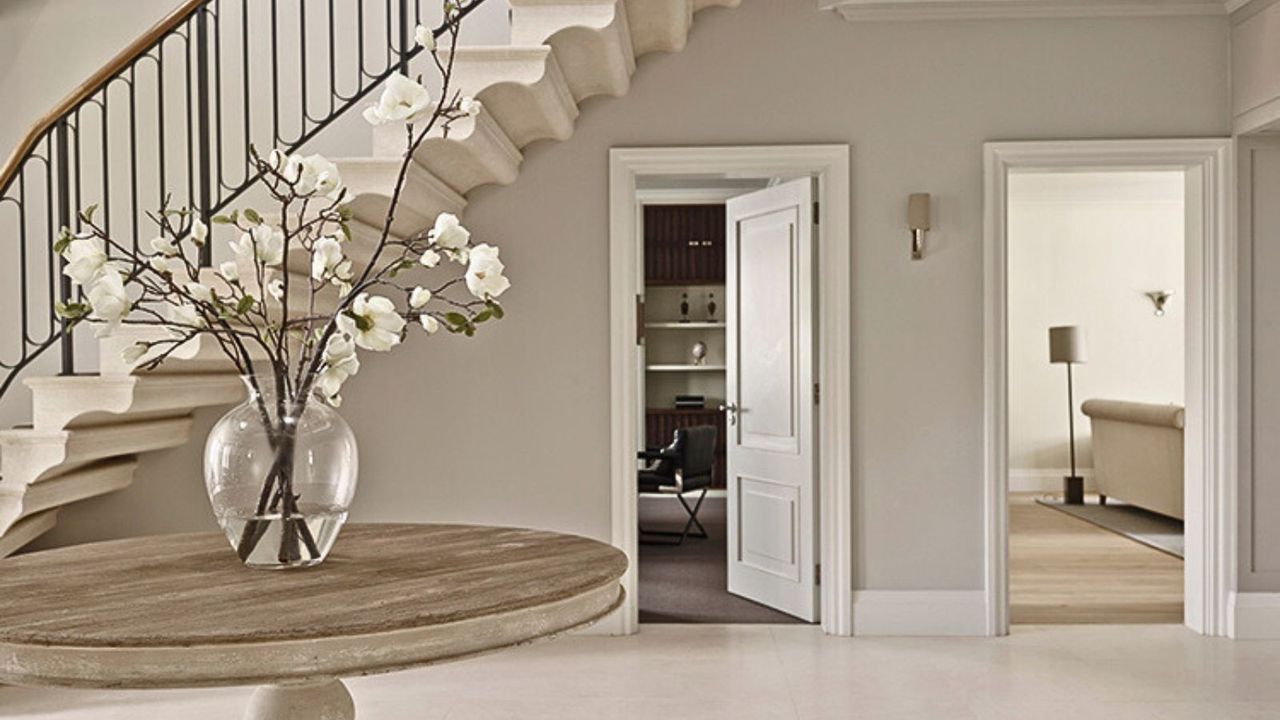 A large neutral colored entryway with a large center table with a vase of orchids, and an open door under the stairs leading to a home office