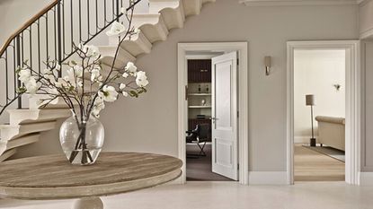 A large neutral colored entryway with a large center table with a vase of orchids, and an open door under the stairs leading to a home office