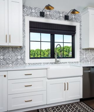 A kitchen with a black window and blinds, black and white patterned walls, and upper and lower white cabinets