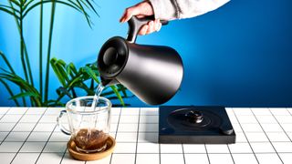 the fellow Corvo EKG kettle in matte black photographed against a blue background with a glass mug and black and white table