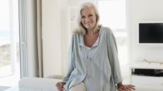 A retired woman sits on her couch smiling.