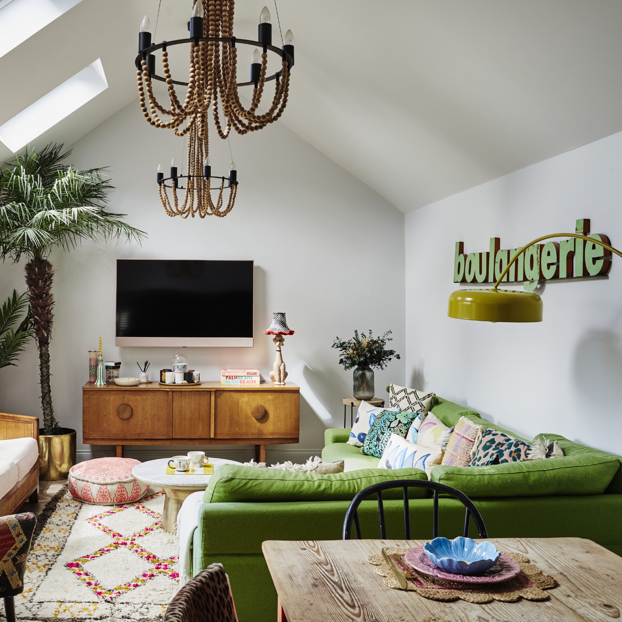 A white living room with a green corner sofa, a 'boulangerie' sign hanging on the wall and two potted palm trees in the corner