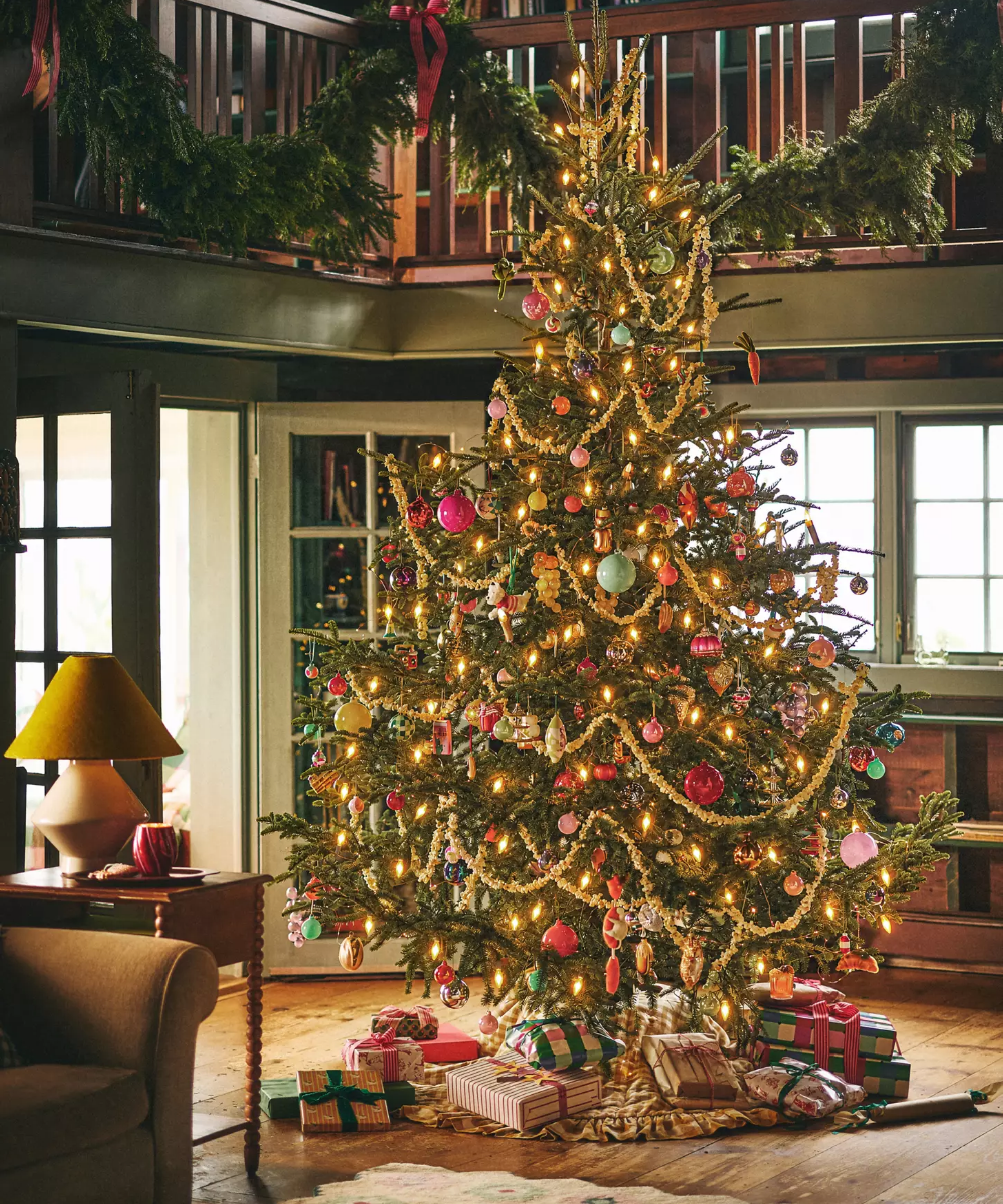 Colorful, eclectically decorated Christmas tree set in the center of a rustic living room surrounded by overflowing wrapped gifts with classic evergreen garland draped across the banisters overhead