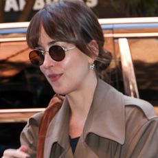 Dakota Johnson wearing a brown trench coat and jeans with a brown suede hobo bag.