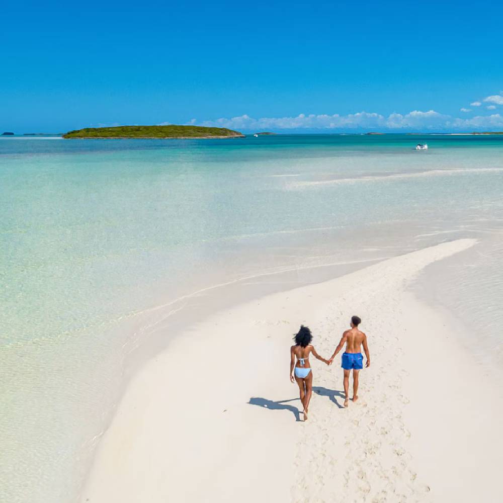 A man and a woman hand in hand, walking across a luxurious beach with blue water in the distance.