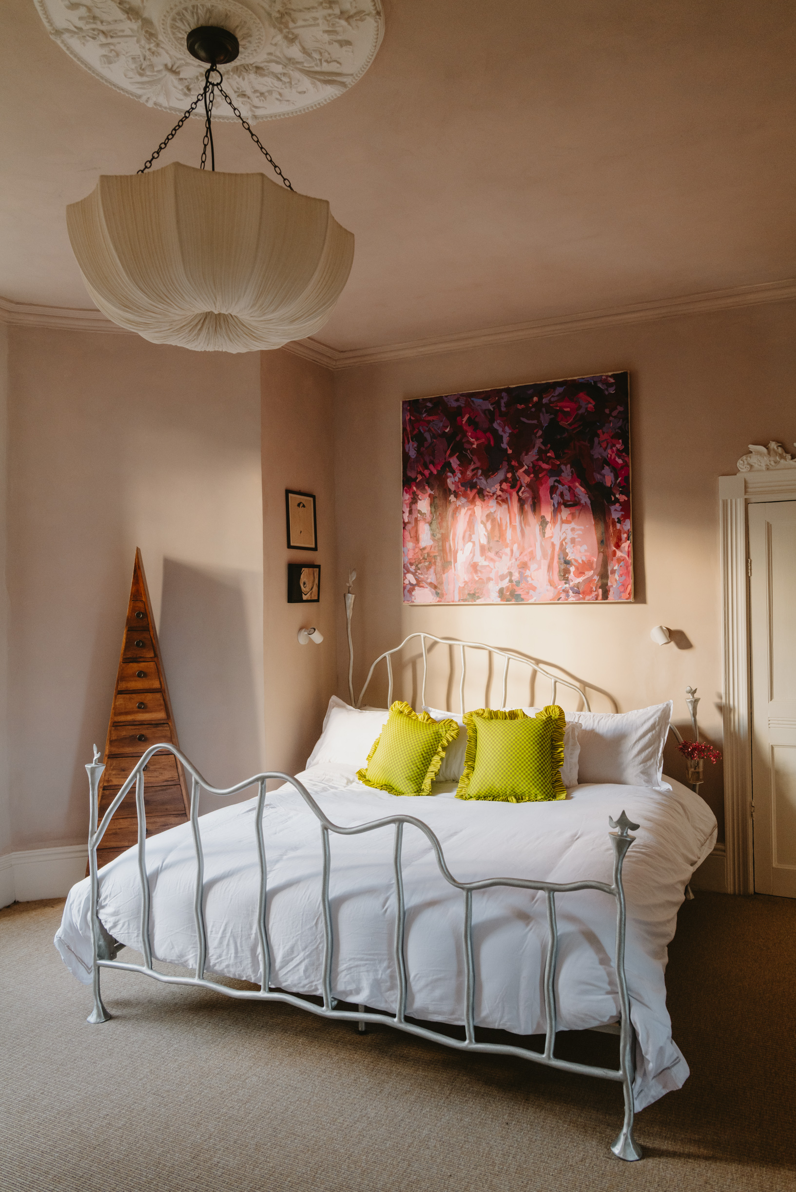 A modern bedroom with an abstract metal bed frame, with white bedding and two lime green scatter cushions with frilled edges, with blush pink on the walls and a large pink patterned artwork