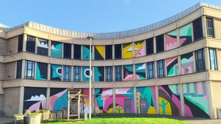 An abstract, brightly colored mural covers the ground floor and second level of a curved, beige-colored building, featuring geometric shapes, cloud forms, and stylized faces.