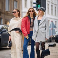 Models pose on the streets of Copenhagen. On wears a kimono and khaki pants. One model wears Black top and red jacket and blue jeans. One wears a ripped suit set and black tights and black handbag. 
