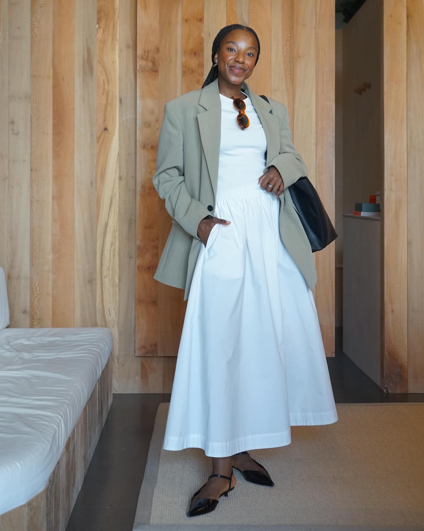 British fashion influencer Taffy Msipa poses at a chic wood-paneled store wearing a neutral blazer, white T-shirt, full white skirt, and black ankle-strap kitten-heels