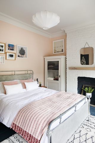 A pale pink bedroom with small white wardrobe, gallery wall overhead, rectangular mirror and disused fireplace