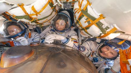 three men in white and blue striped pressure suits are seen from above inside a cramped space capsule