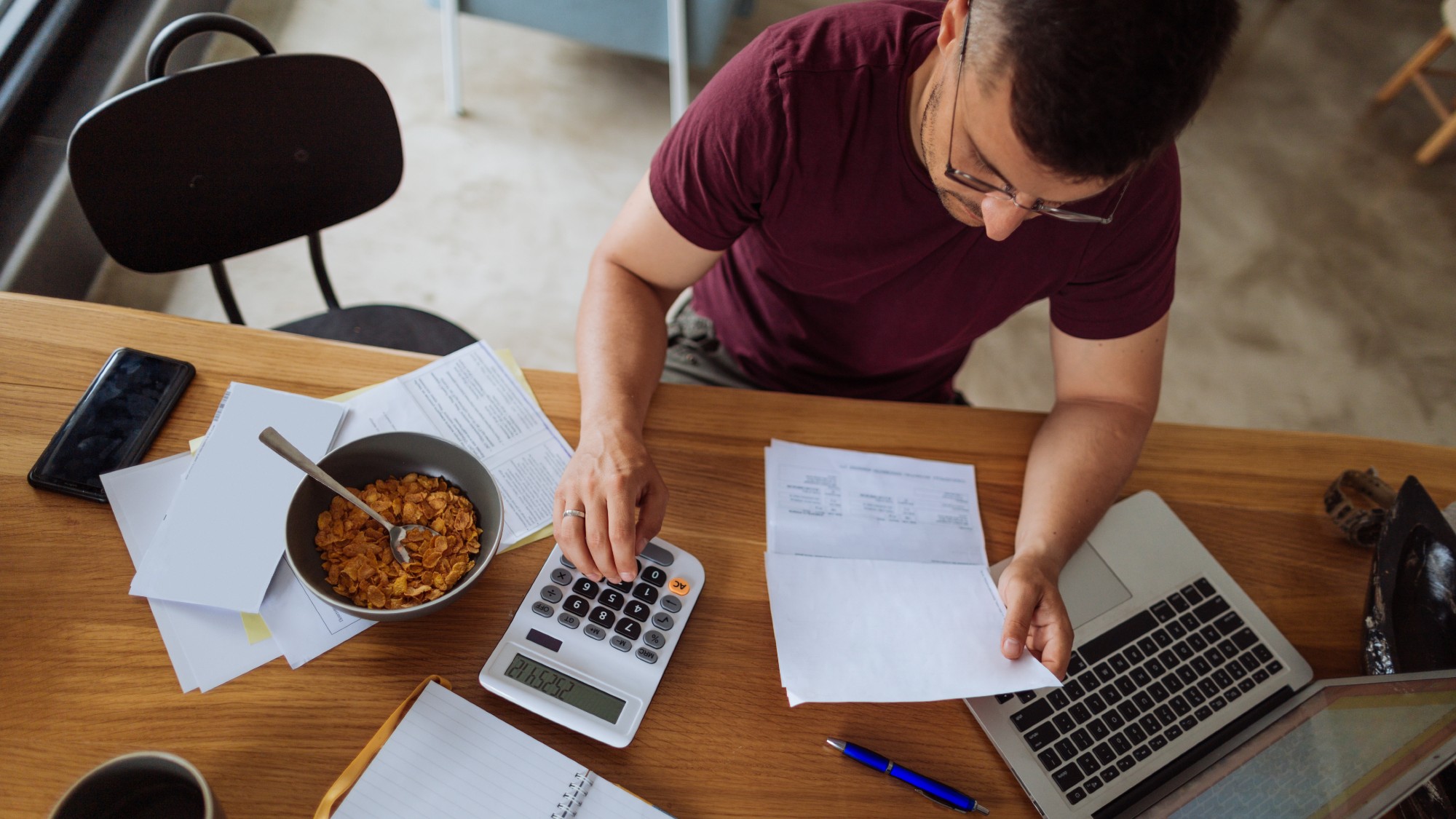 Gig worker sitting at a desk at home paying bills with a calculator, laptop, and bowl of cereal