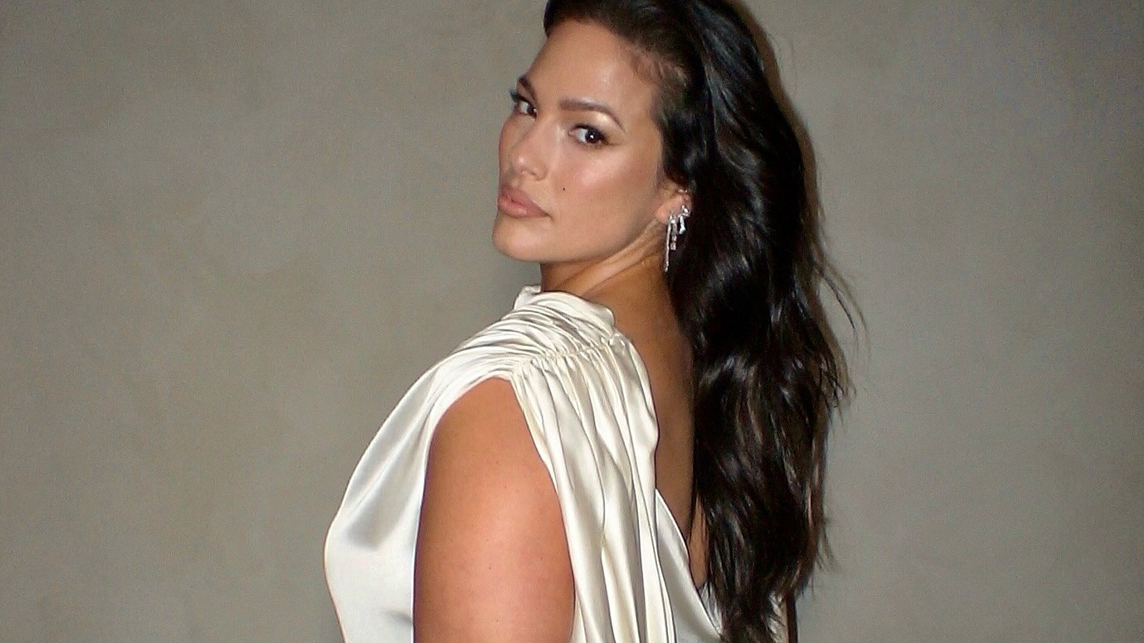 Ashely Graham wearing white plus-size formal dress while standing against gray backdrop.