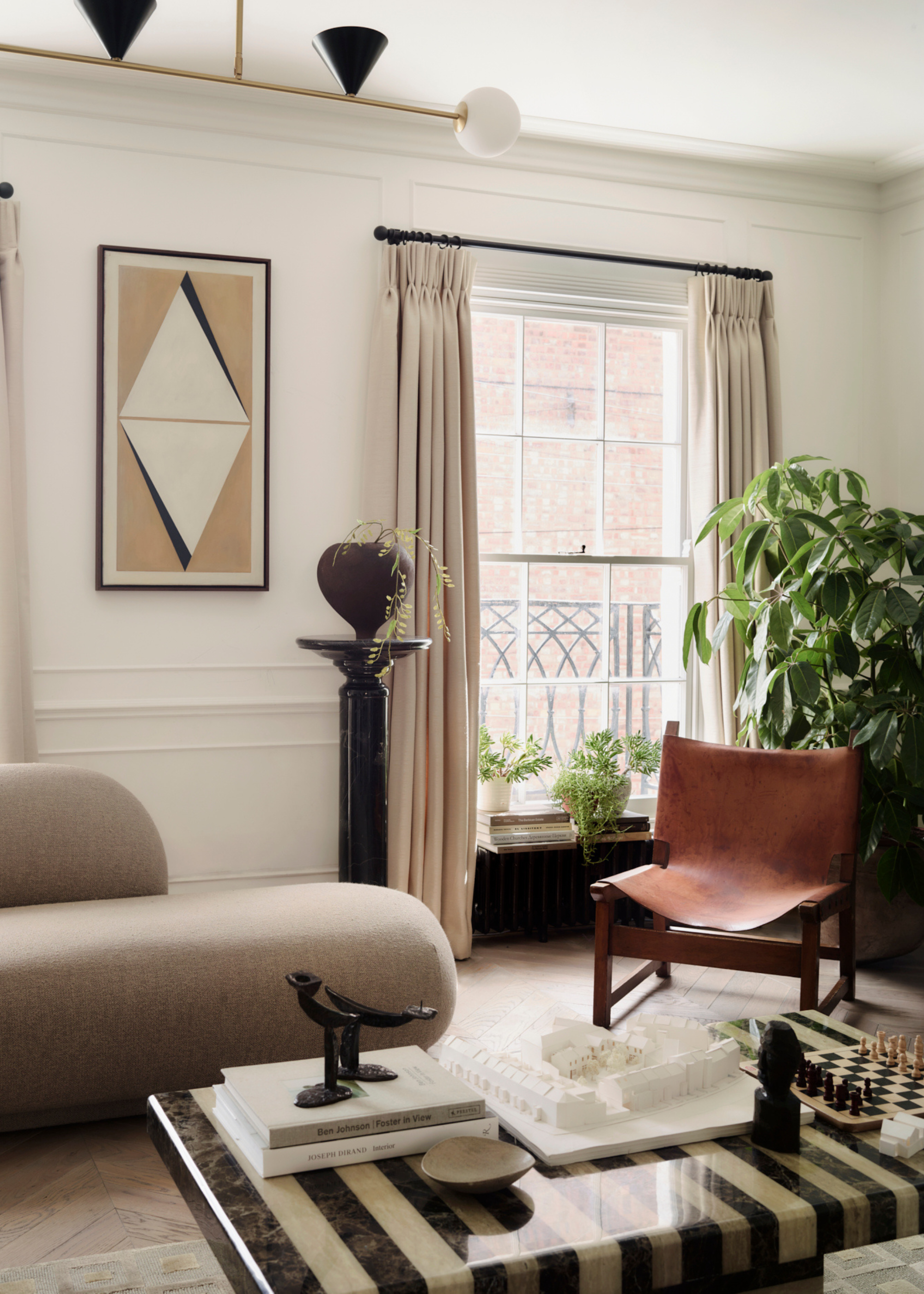 A minimalist living room with a beige sofa, a leather armchair, a vase on a pedestal, potted plants, and a mixed marble side table with books, candle holders, a chess board, and a ceramic bowl