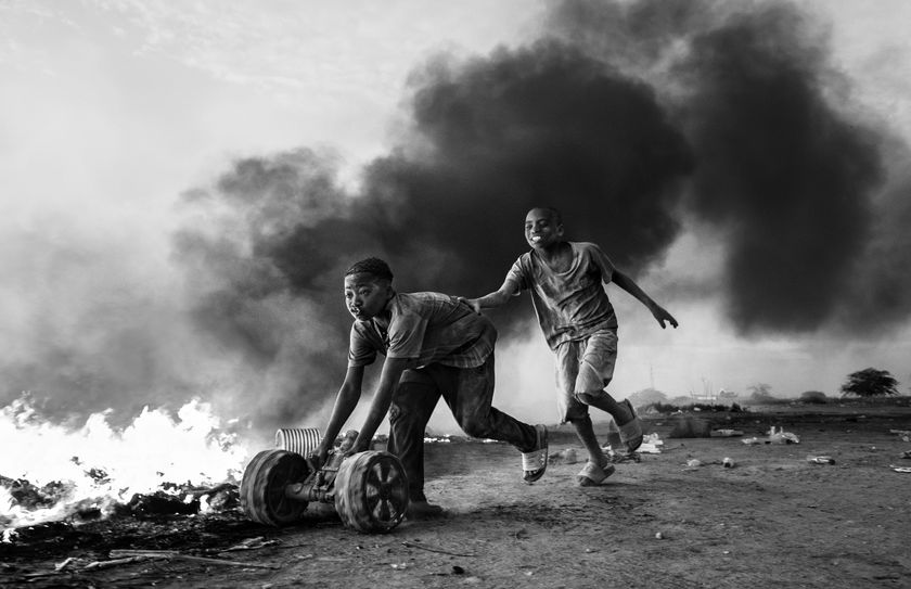 Two individuals run away from a blaze, pushing a cart amidst thick black smoke and a chaotic, desolate landscape