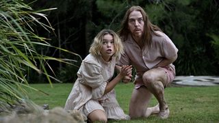 Milly Alcock in a cream dress as Meg and Tim Minchin in pink pyjamas as Lucky crouch on a lawn looking anxious in Upright