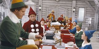 Will Ferrell sitting at a table with elves and looking gigantic.