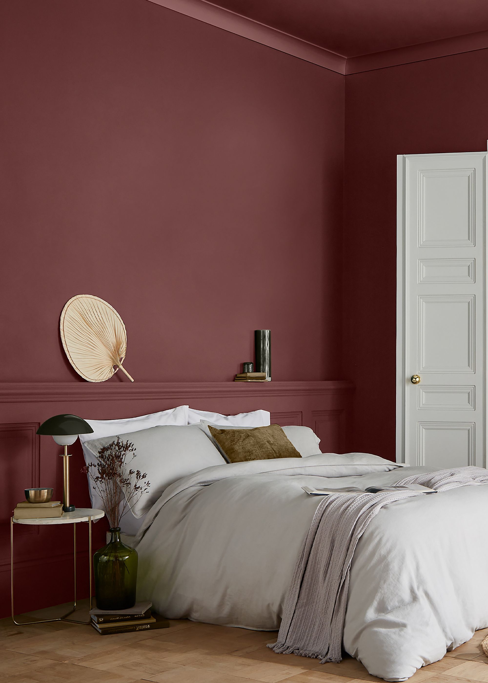 A modern bedroom with burgundy color-drenched walls, wood flooring, light gray bedding, decorative wall panelling, and a white door with paneled detailing.