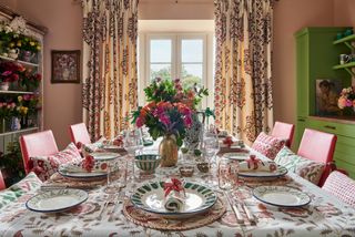 colorful dining room with pink walls, floral print drapes, floral tablecloth and napkins, patterned plates, bright red chairs, shelving unit full of flowers, green kitchen cabinetry on right