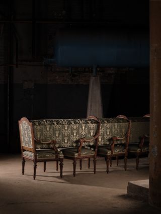 A chair sculpture. Chairs are set one behind the other, with textile objects going through each back of a chair, in continuation. It's set in an industrial factory setting.