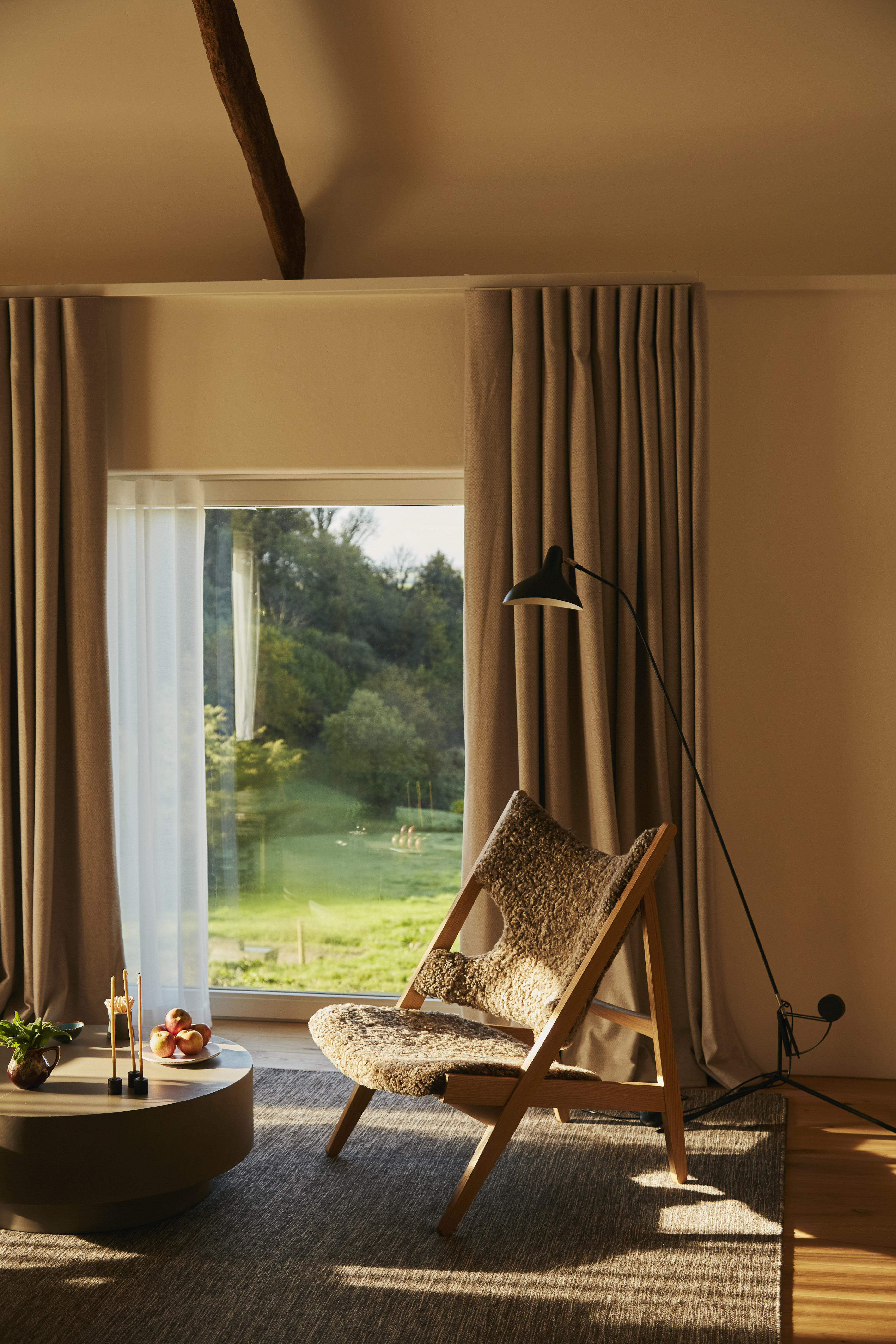A hotel room at sunset features a goldeny natural light filtering through floor-to-ceiling windows, modern rustic decor, including wooden floors and furniture, a neutral rug, and fruit, candles, and plants on display.