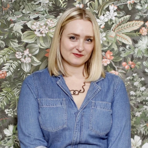 Image of a woman in a denim shirt standing in front of a floral patterned wallpaper