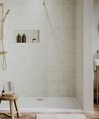 A white tiled bathroom with an open shower with a gold sink and built-in shelves, with a circular wooden stool and white textured bath mat