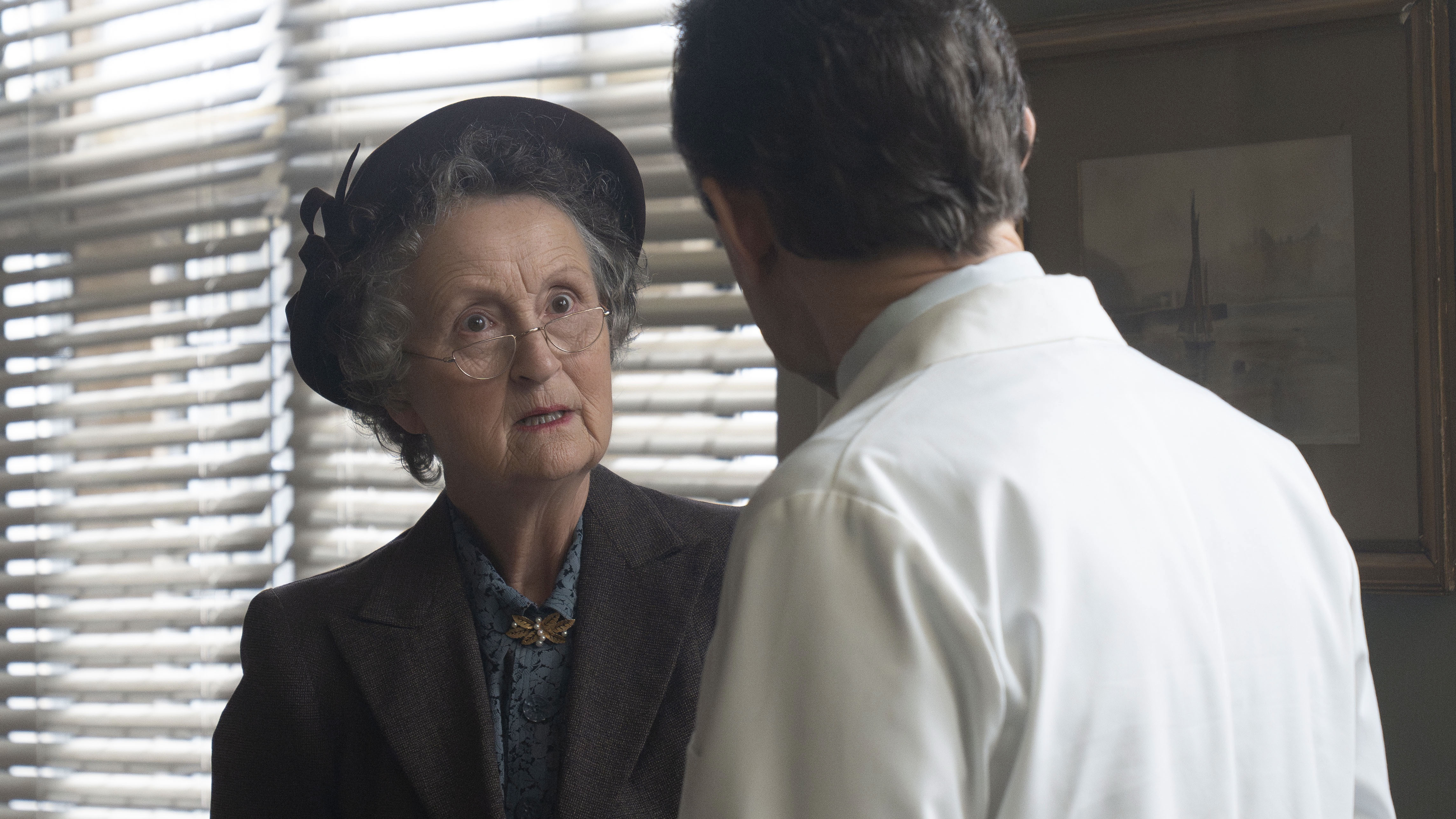 Georgie Glen in a dark suit and hat and glasses as Miss Higgins talks to Stephen McGann in a white coat as Dr Turner in Call the Midwife.