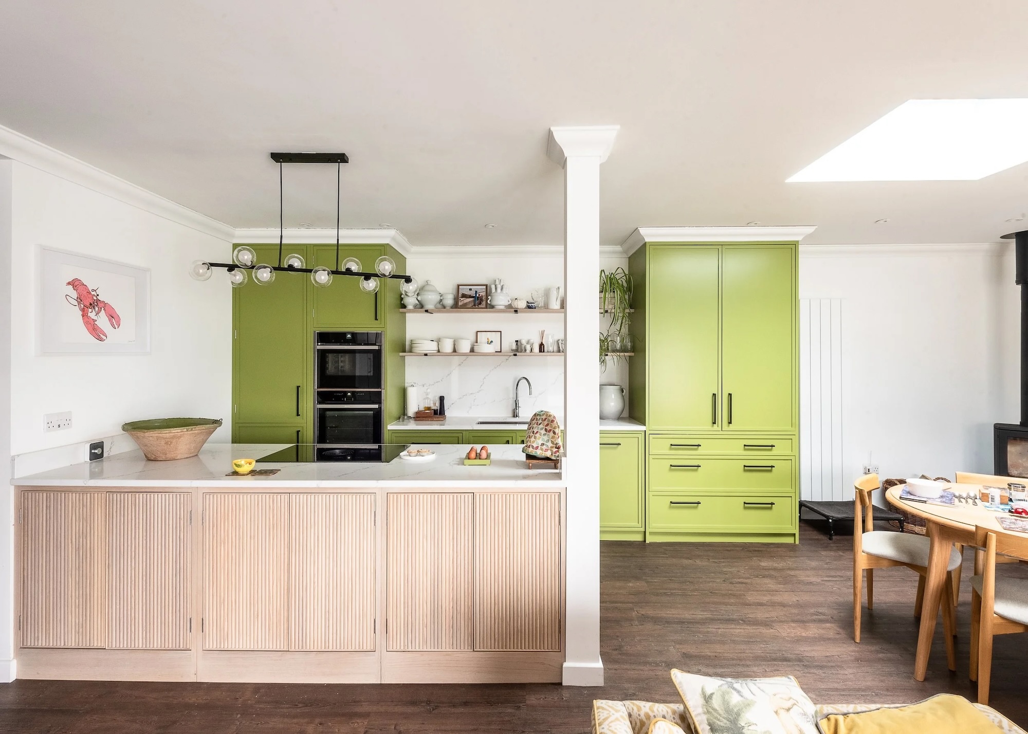 A light oak and green colored kitchen with large green cabinets and doors as well as an island with a stove on top. There is also a view of the dining area.