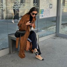 Woman wears Adidas track pants, a Dior saddle bag, Chanel flats and a brown trench coat while texting on the street. 