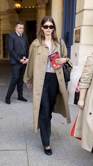Kaia Gerber wearing a trench coat with a grey sweater, trousers, and black flats
