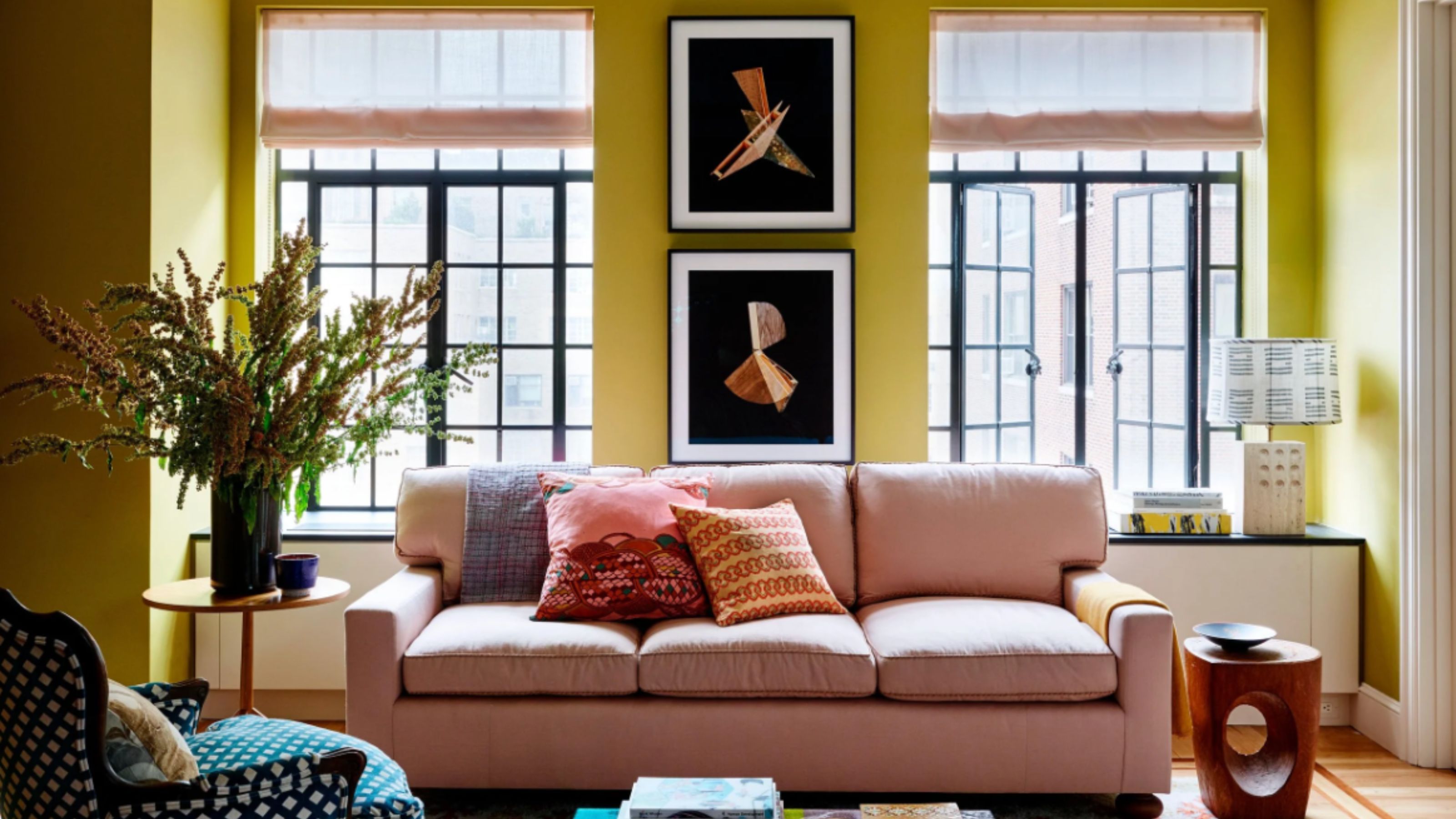A modern living room with chartreuse walls, a white ceiling, two large windows, a pink sofa with colorful cushions and a modern multi-colored glass coffee table. 