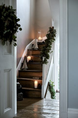 A hallway with wooden floorboards, white painted Staircase with candle lighting in hurricane vases and greenery on banister
