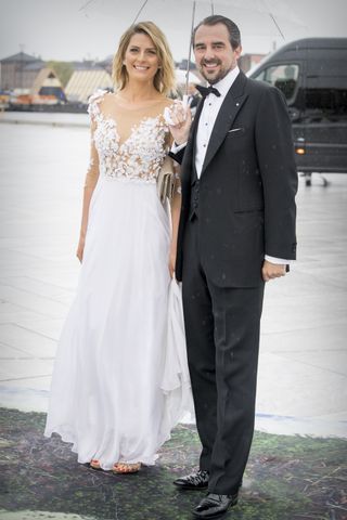 Princess Tatiana wearing a white gown and Princess Nikolaos wearing a suit standing under a clear umbrella standing in front of a black van