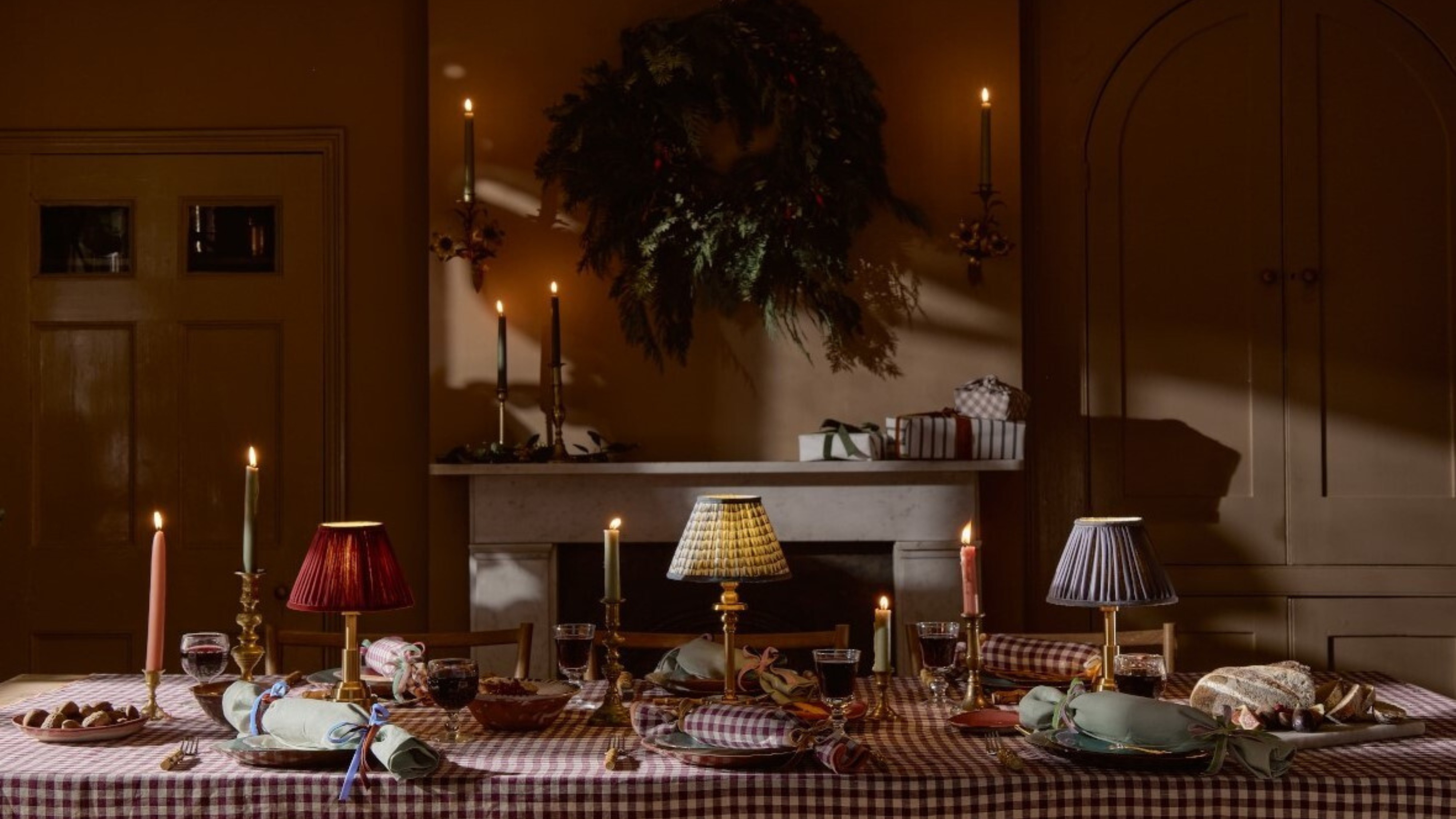 Moody extra-long holiday dining table styled with gingham linens, taper candles, and portable lamps.