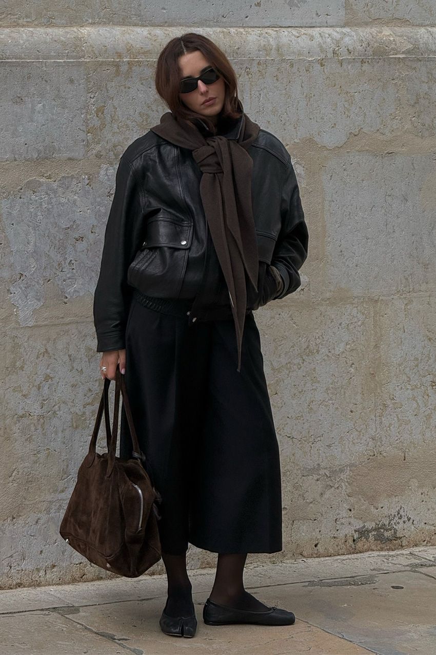 A woman standing outside in front of a concrete wall wearing a large black bomber jacket with a black midi-skirt, black tights, black flats, a brown suede bowler bag, black sunglasses, and a brown scarf tied around the shoulders.