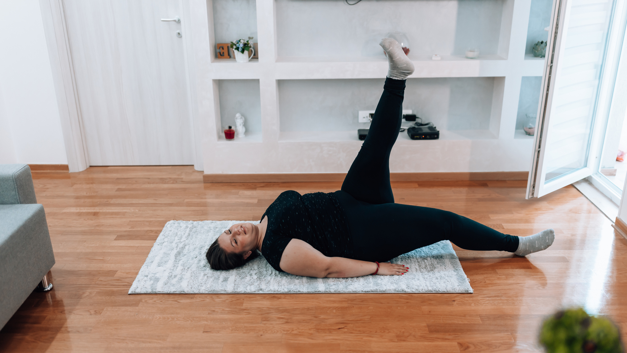 Woman wearing a black top and black leggings lies on her back with one raised in a living room