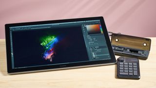 The Huion Kamvas Pro 19 featuring artwork of recolored fireworks, next to the the stylus case and Huion Keydial Mini shortcut remote, in front of a pink background.