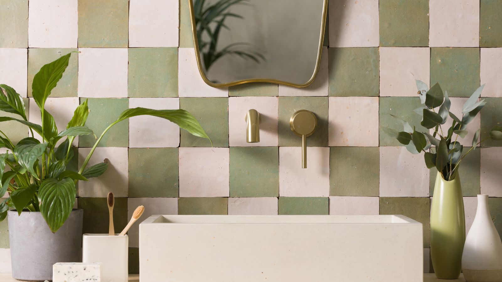 A white and green Zelige tiled backsplash wall with a white sink, gold faucets, and a gold mirror above. To either side are green houseplants.
