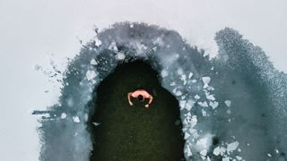 Unidentified man in a lake, in ice-hole of icy water covered with white snow and ice.