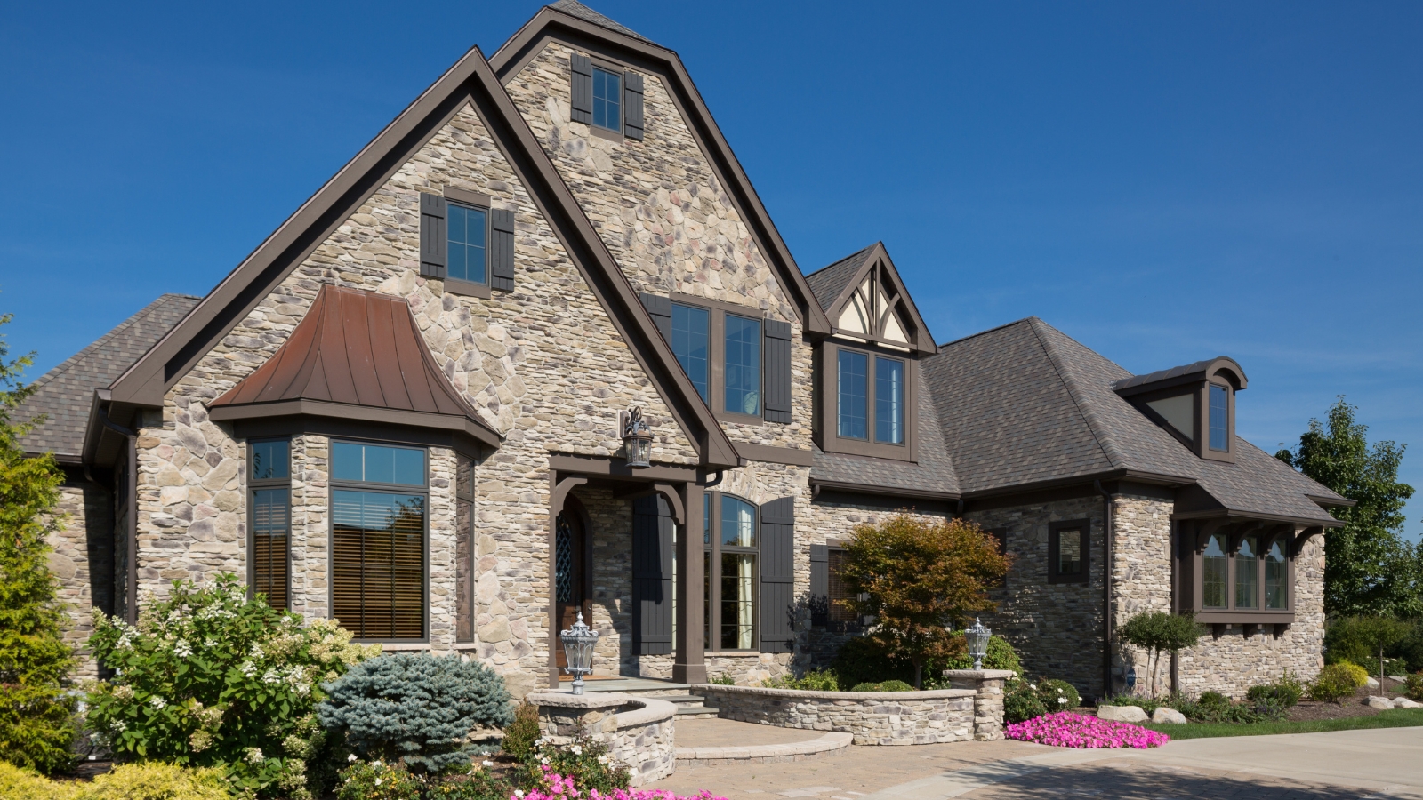 Exterior of large, affluent, stone house with arched roofs and windows. Foliage and colorful flowers are pictured in front of the house, and there is a blue sky.