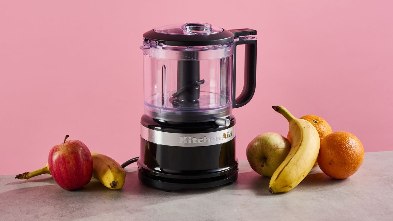 A black gloss KitchenAid 3.5-cup mini food chopper sits on a grey stone-effect surface. The background is pink, and there are apples, bananas and pears on the surface around the chopper.