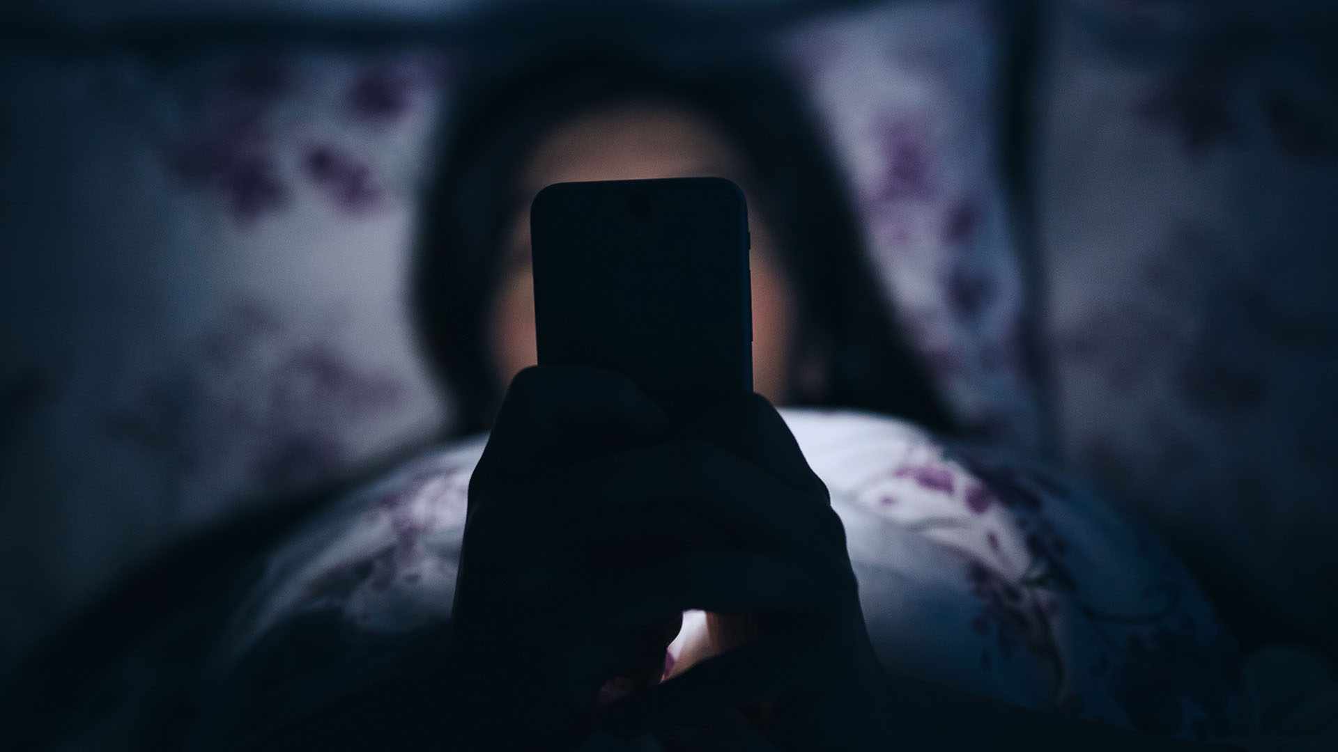 A woman in bed holds a phone in front of her, the screen light on her face