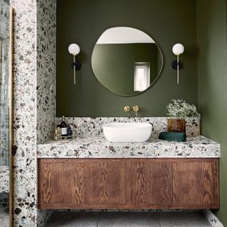 a green bathroom with a vanity area with a single white basin, terrazzo wall and countertop, wooden floating vanity unit, globe wall lights and round wall mirror