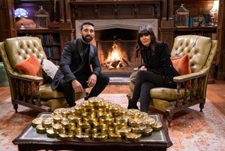 Jaz sits in an armchair in the Traitors castle opposite Claudia Winkleman in a separate armchair. There is a roaring fire behind them, and in the front of the shot is a coffee table with a sprawling pile of gold coins on it.