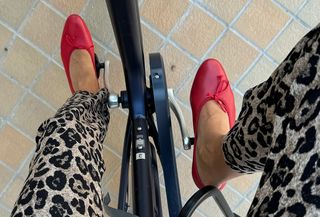 Anne wears leopard pants with red ballet pumps while riding a bike.